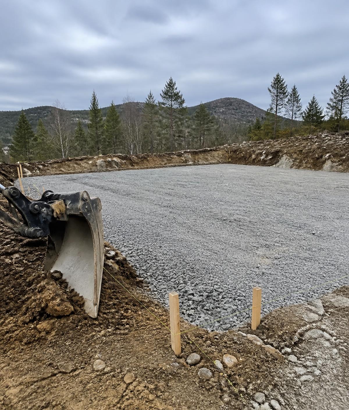 Illustrasjonsbilde: planert tomt med pukkdekke og gravemaskinskuffe i forgrunnen.